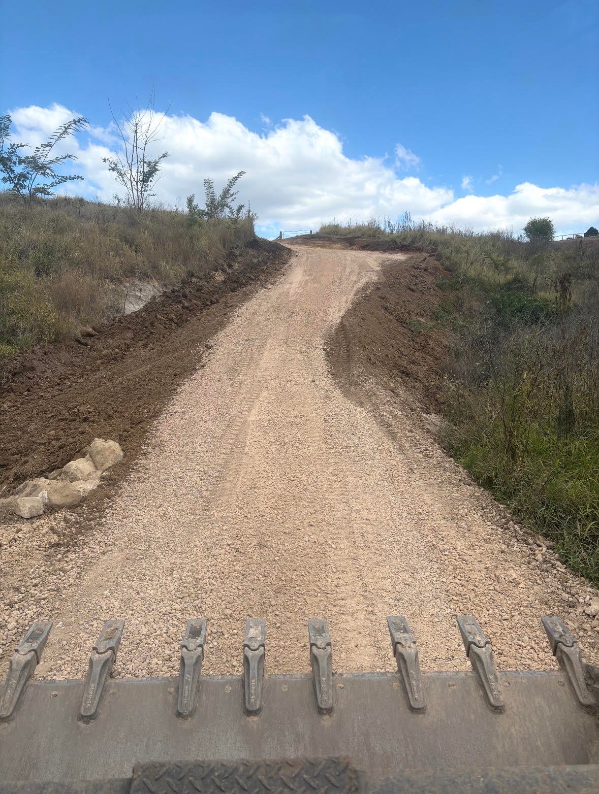 Unpaved gravel road stretching through rural landscape with sparse vegetation under blue sky with clouds