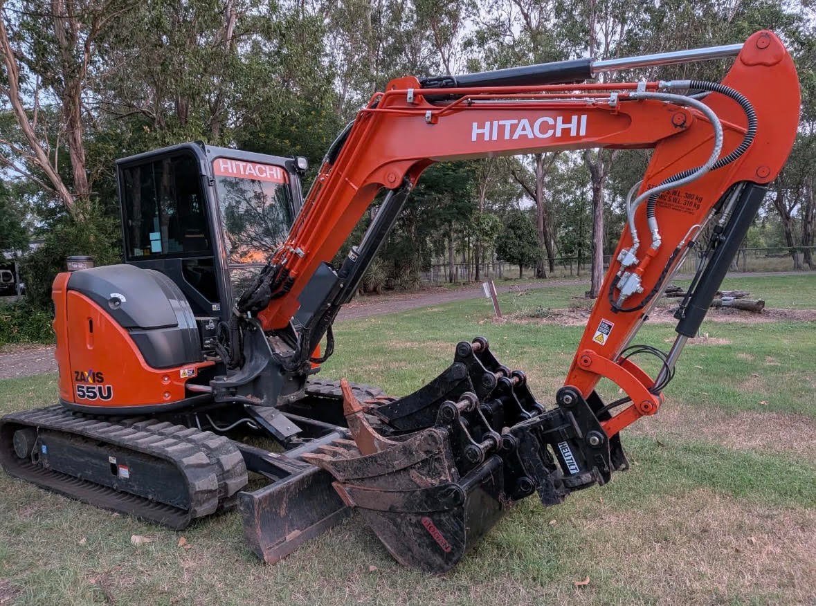 Red Hitachi mini excavator with extended boom and bucket attachment on grassy terrain with trees