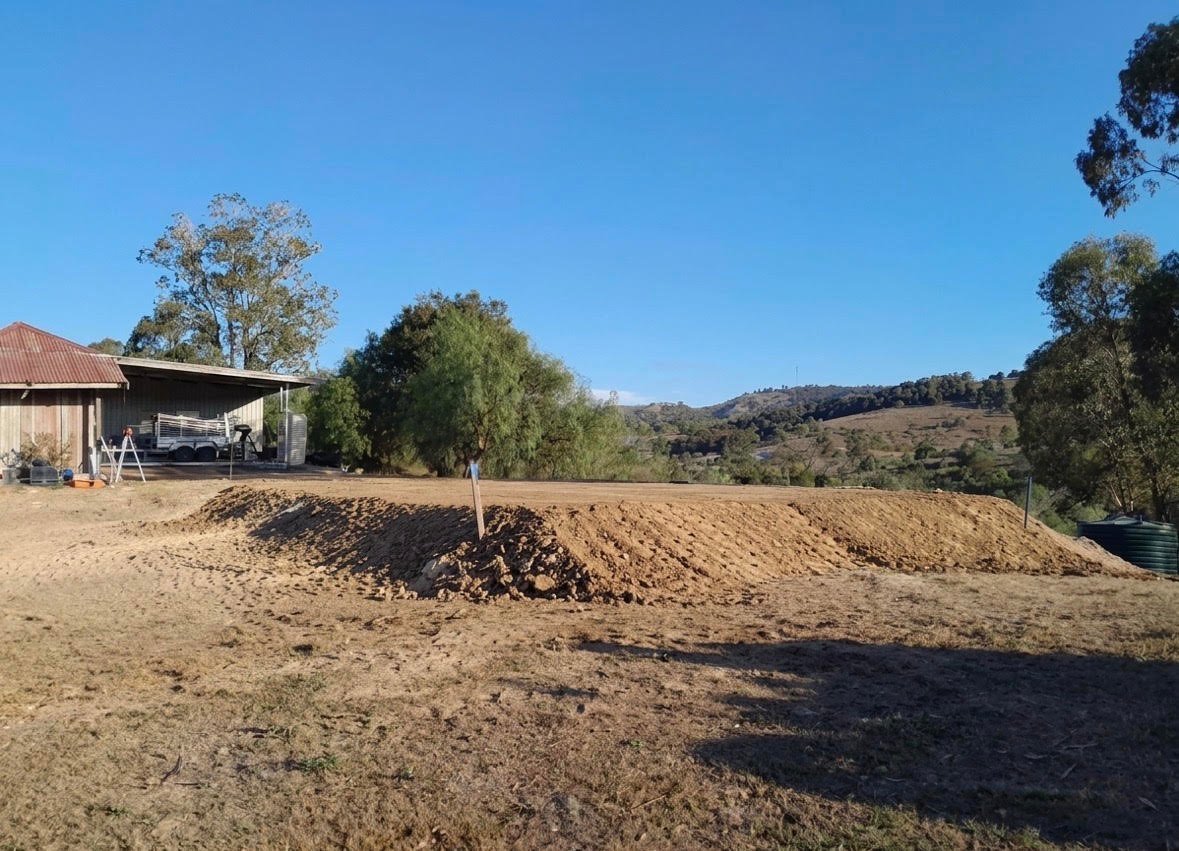 Rural property with farm shed, dirt pile, trees, and hillside landscape under clear blue sky