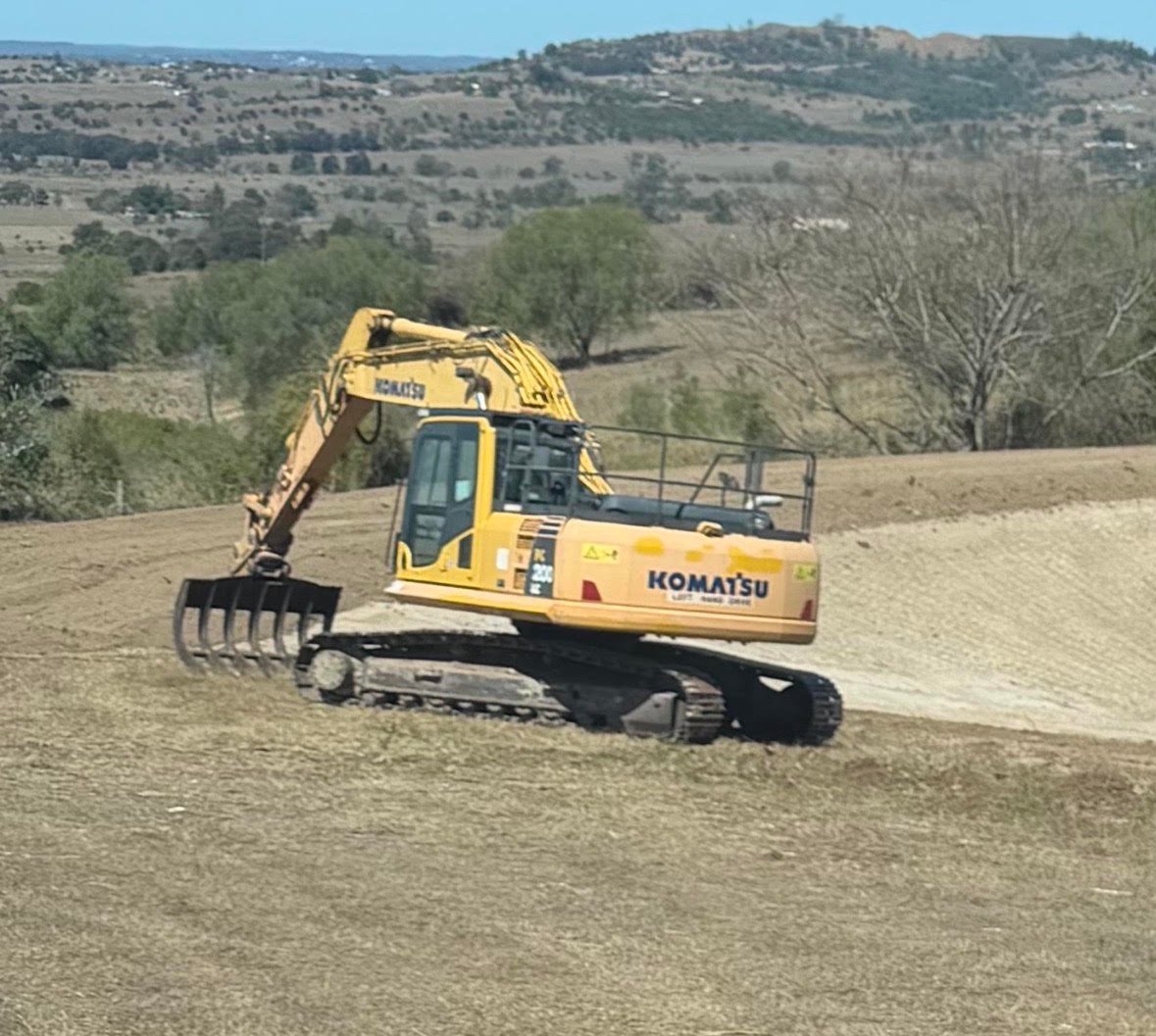 Yellow Komatsu excavator operating on a hillside with rural landscape and trees visible in the background